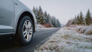 Voorwiel van een zilvergrijze auto op een asfaltweg langs berijpt gras en dennenbomen op een koude winterochtend